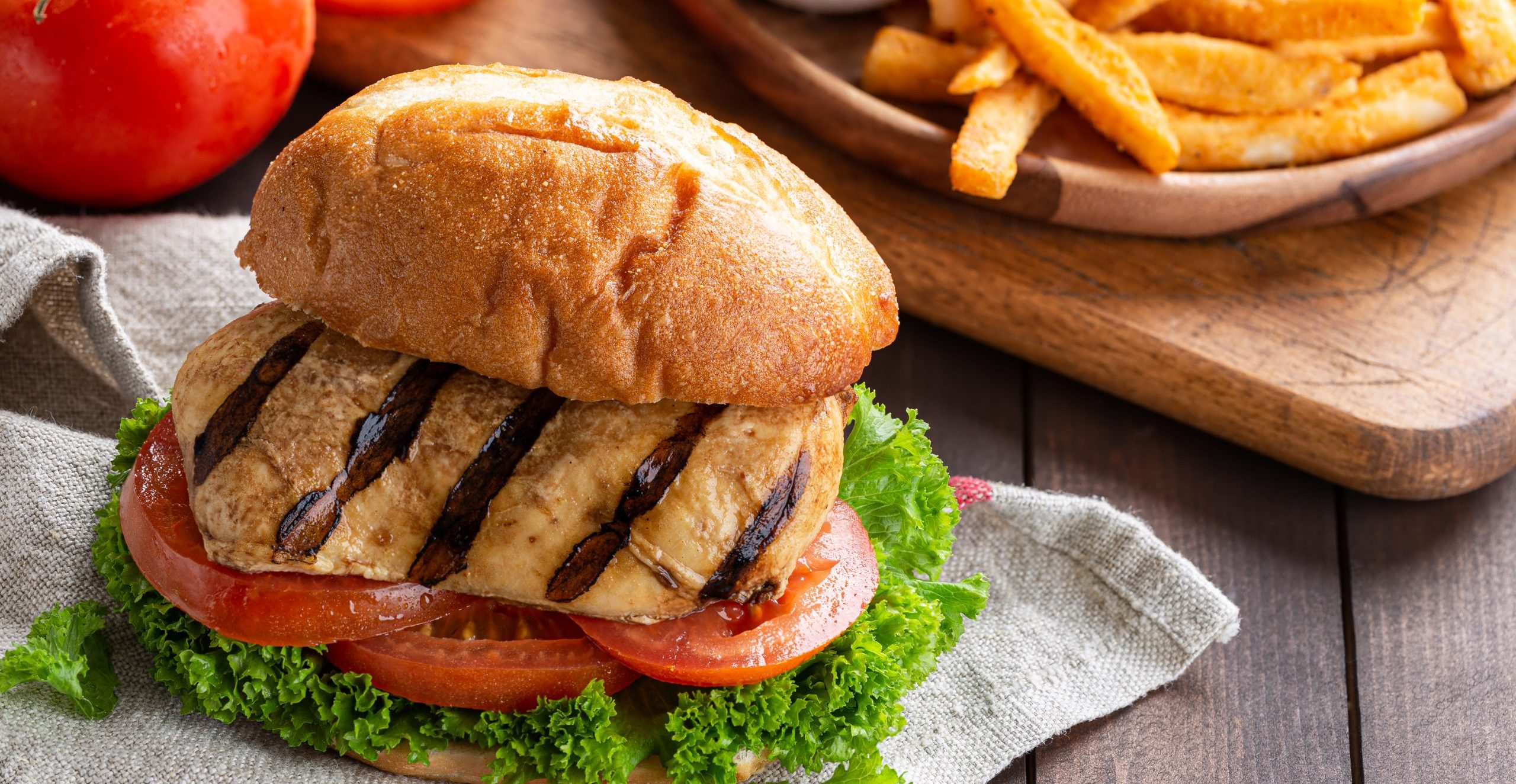 Closeup of a healthy grilled chicken breast sandwich with tomatoes and lettuce on a bun with french fries in background